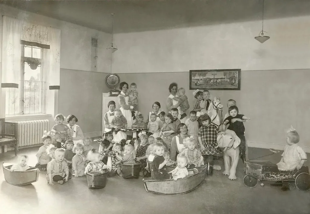 Svartvitt foto av många barn och kvinnor i en förskola med leksaker som små båtar, svampbord, gunghäst och leksaksbil