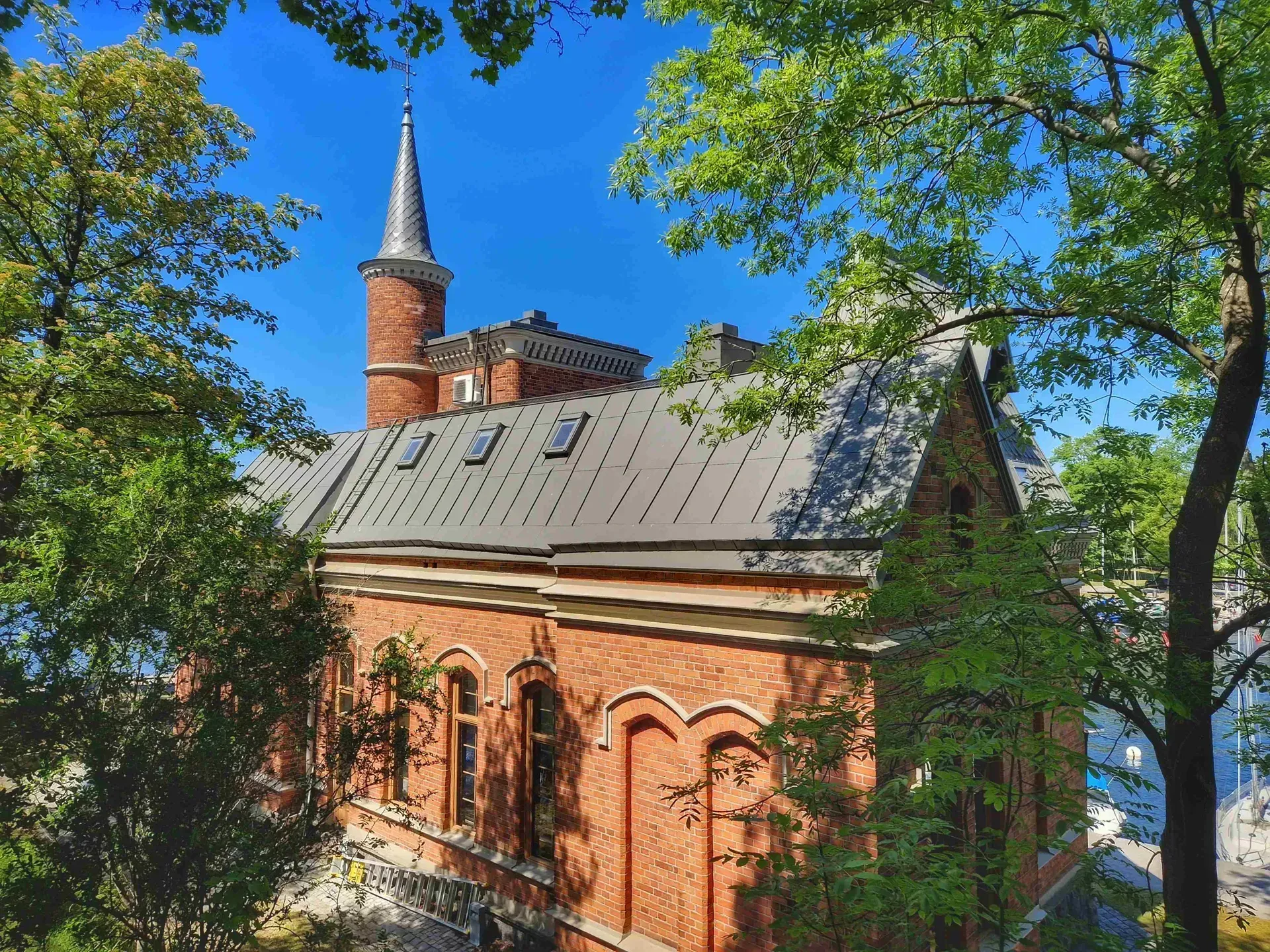 Rött tegelhus med torn, sett genom gröna träd. Grått tak och klarblå himmel
