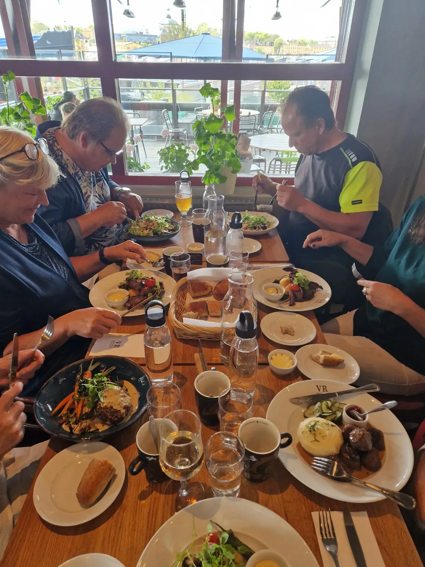 Personer äter lunch vid ett bord fullt med mat och dryck. Fönster i bakgrunden