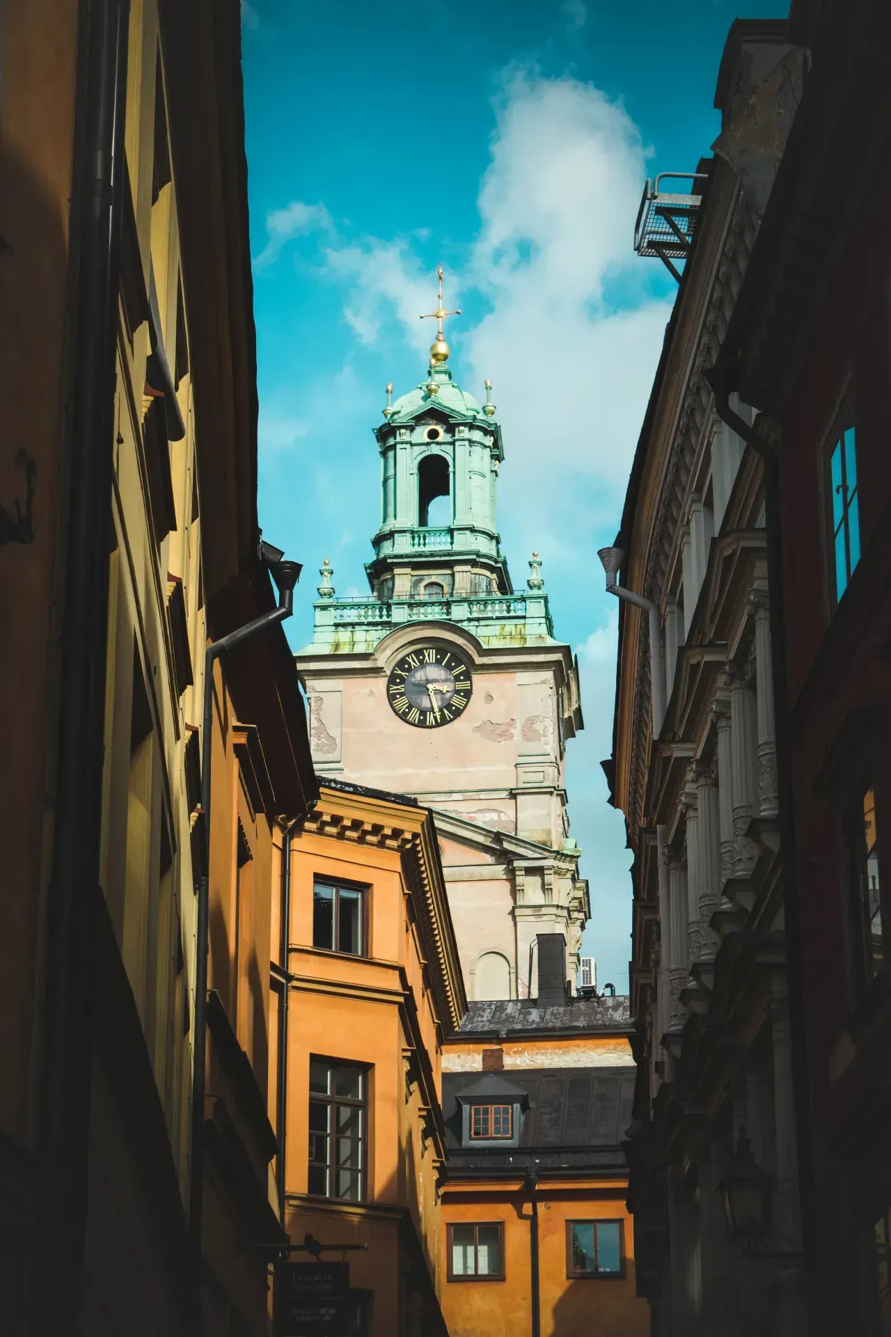 Kyrktorn med klocka, sedd mellan husfasader i Gamla Stan, Stockholm. Blå himmel