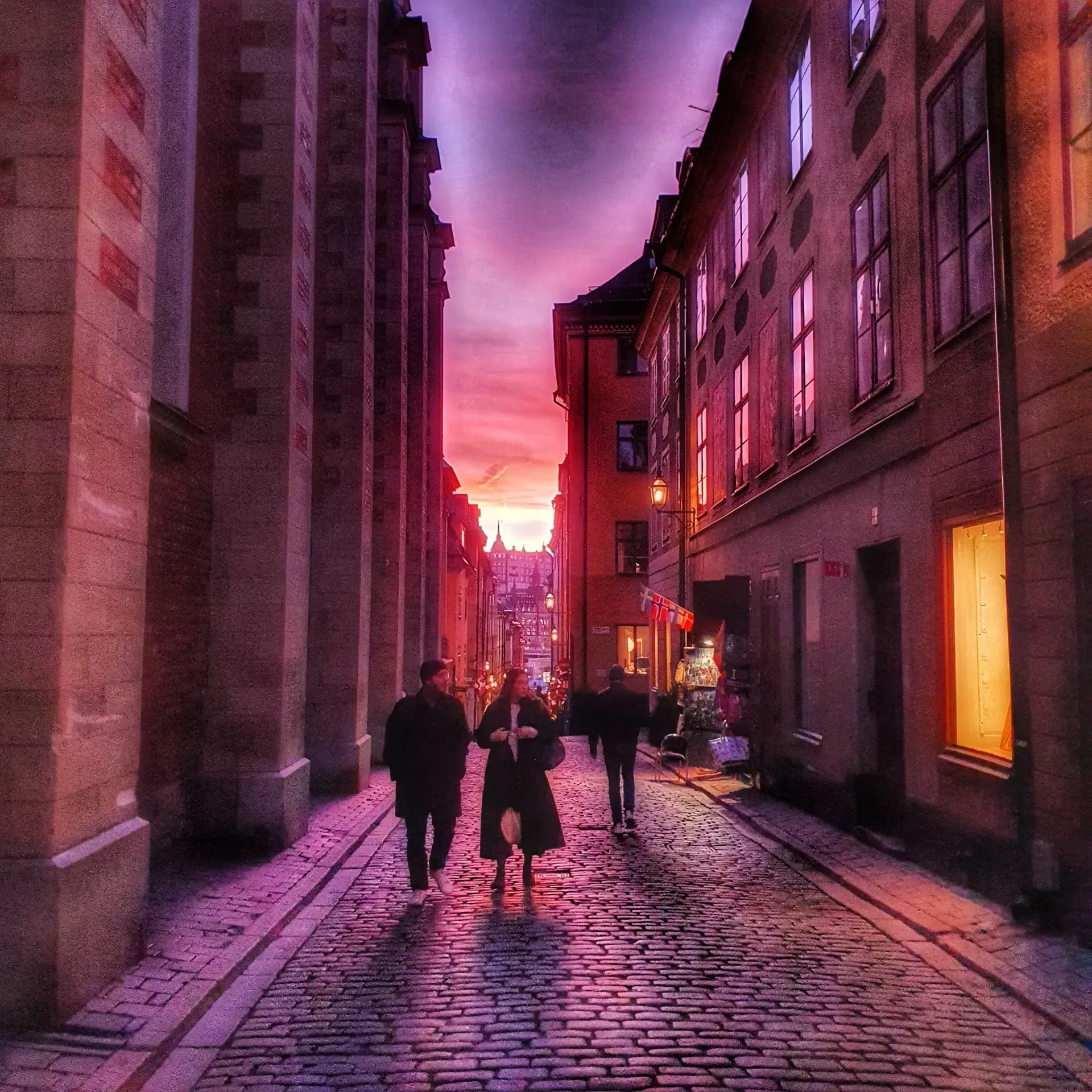 Gata i Gamla Stan med kullersten. Byggnader på sidorna. Folk promenerar. Solnedgång i fonden