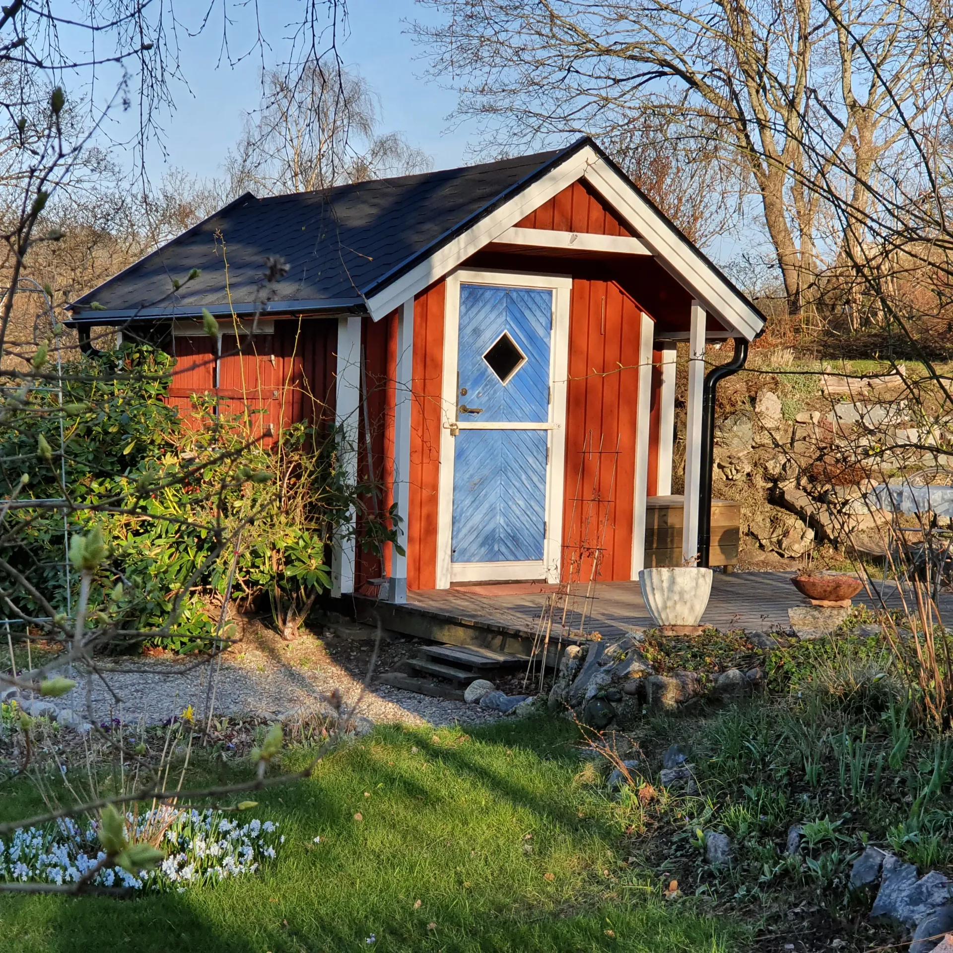 Rött litet hus med blå dörr och svart tak. Trädäck och grönska runt huset