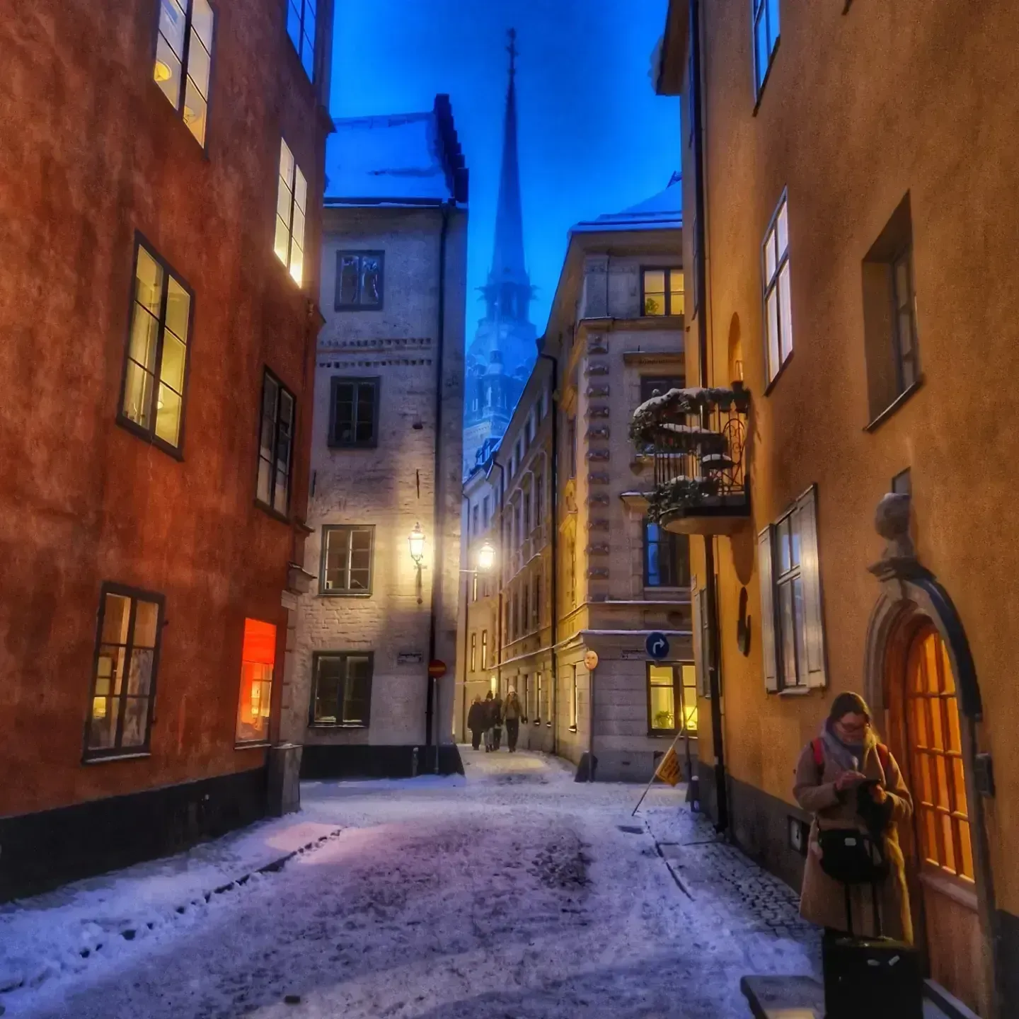 Snöig gata i Gamla Stan, Stockholm, med kyrktorn i fonden. En kvinna med resväska står vid en dörr