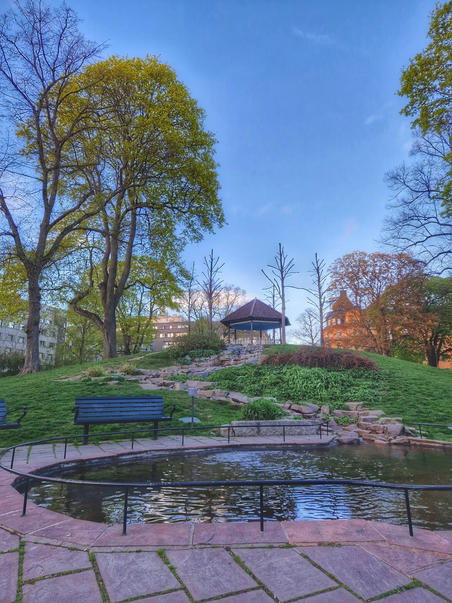Park med damm, bänkar och lusthus på kulle. Träd med gröna löv. Stadsvandring Södermalm