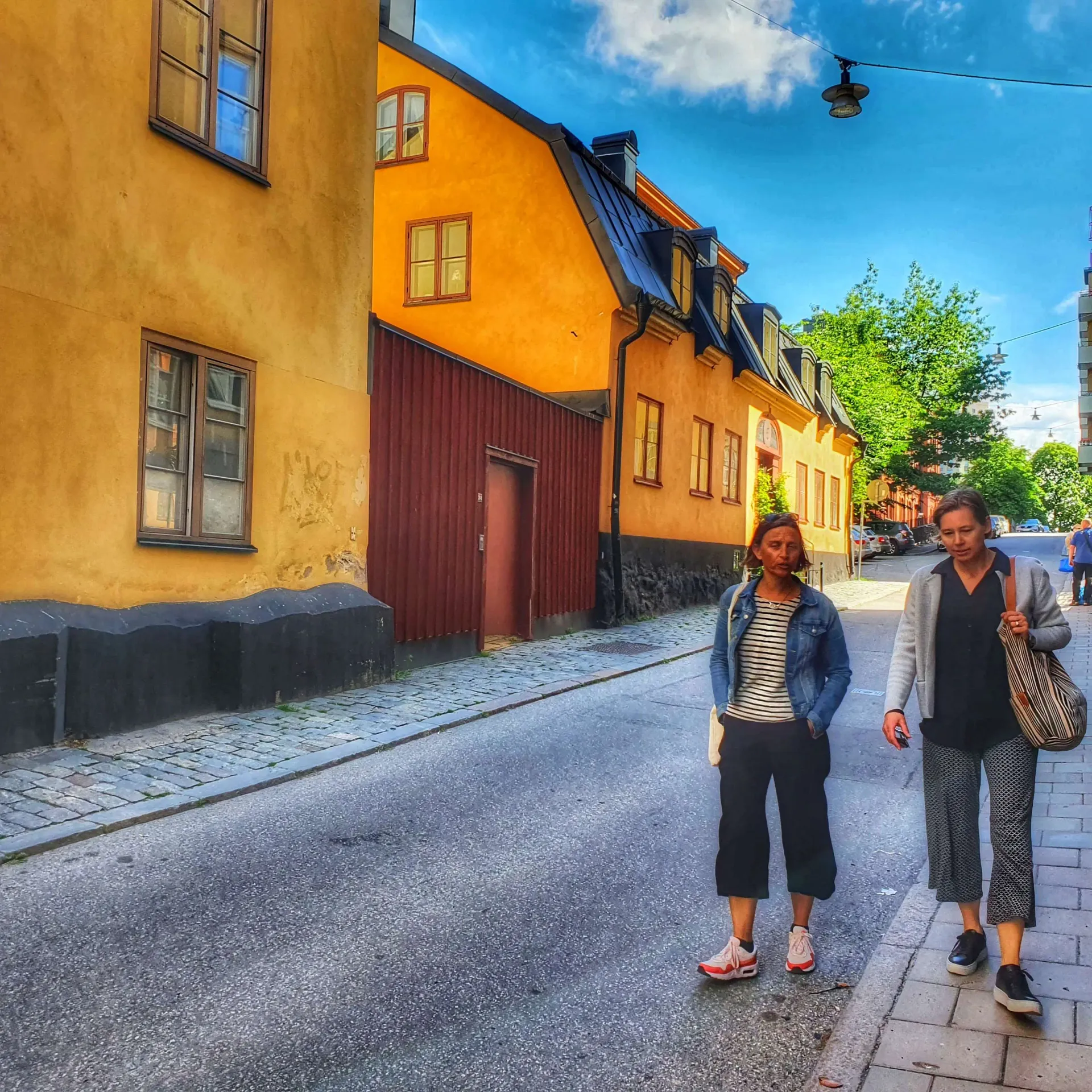 Två kvinnor på gata med gula hus. Blå himmel