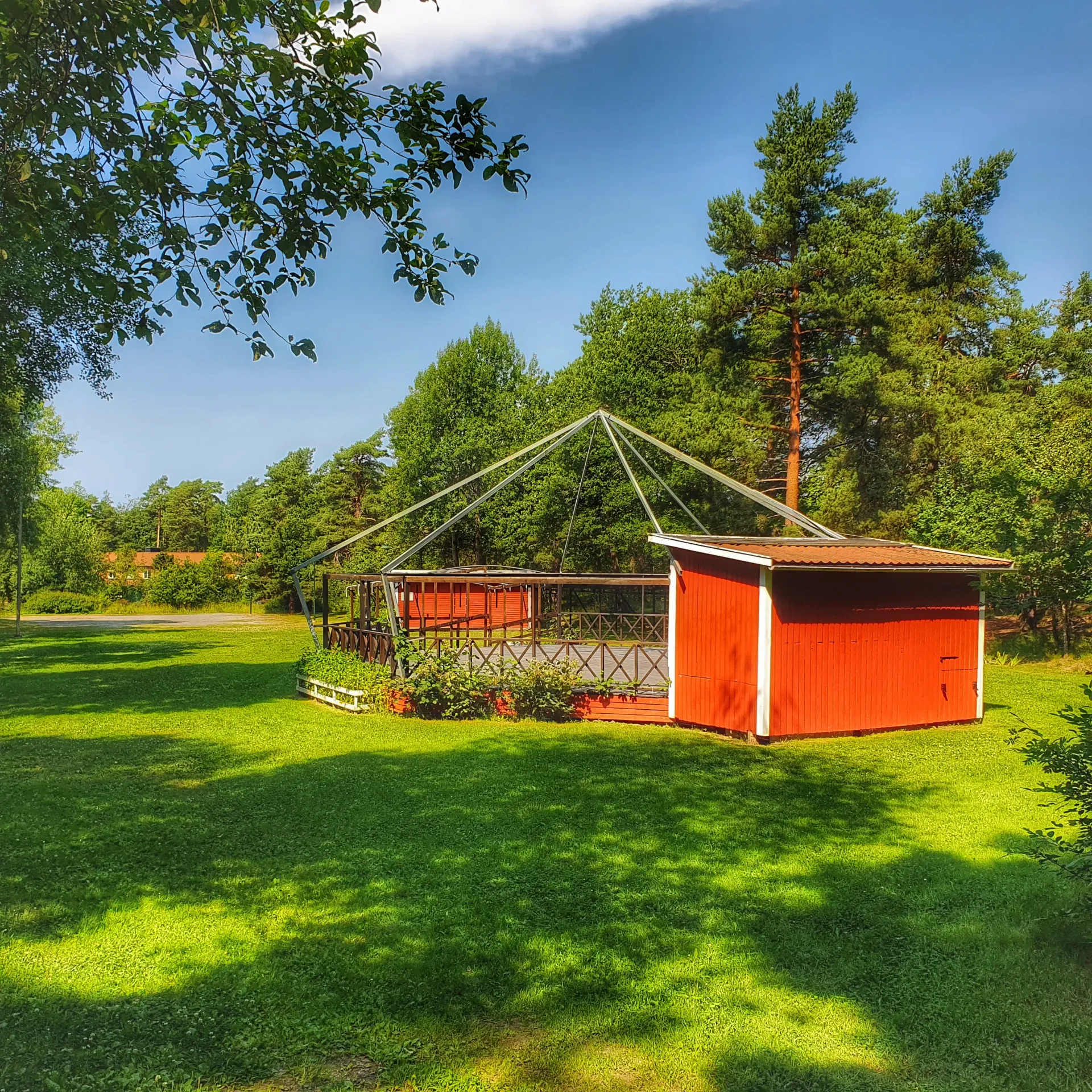 Röd byggnad med trätak och ett tillhörande trädäck i en grönskande park