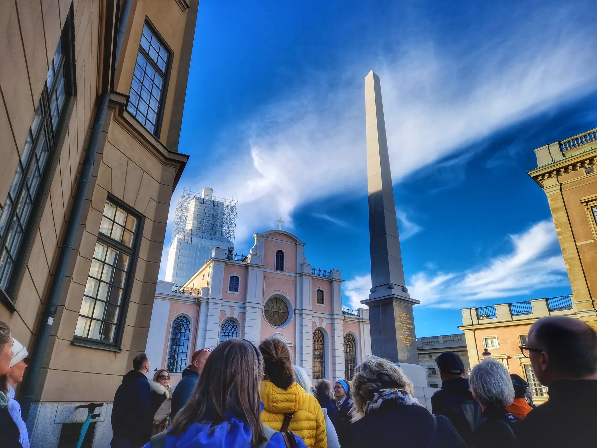 Människor ser mot en obelisk och en rosa byggnad under blå himmel med vita moln. En byggnad har byggnadsställning