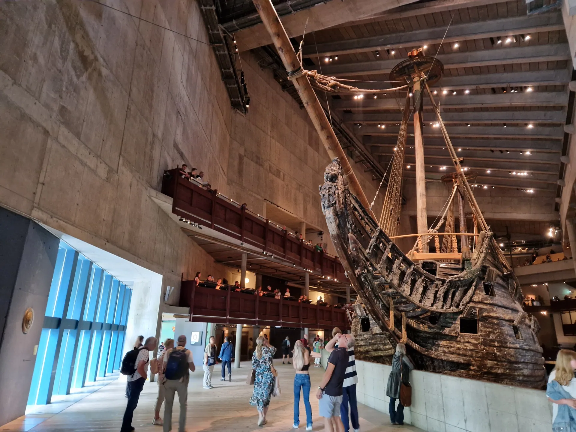 Stort gammalt träskepp i en museihall. Besökare står på golvet och balkonger runt skeppet. Blått fönster till vänster