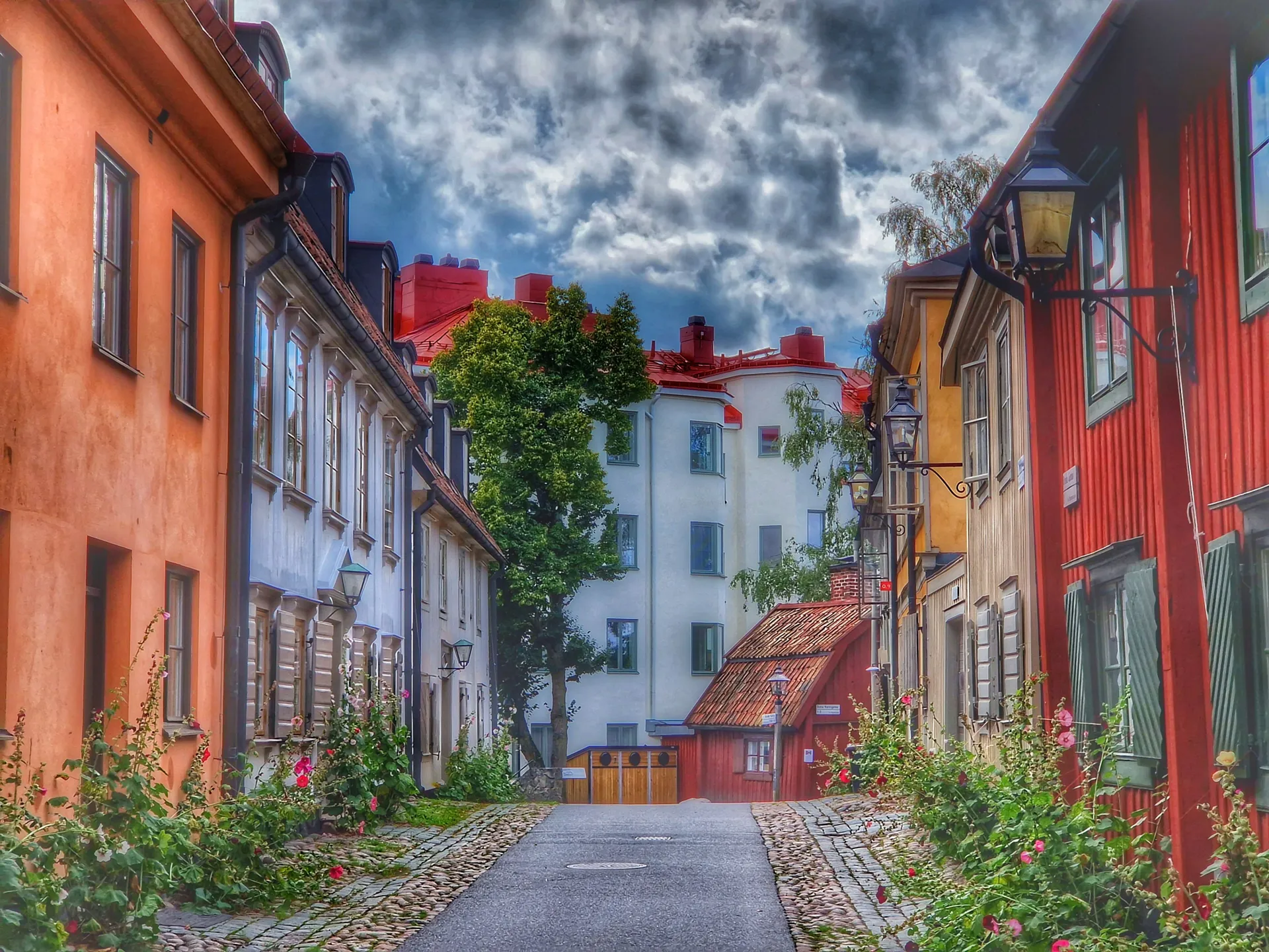 En smal kullerstensgata med färgglada hus, blommor längs väggarna och en molnig himmel