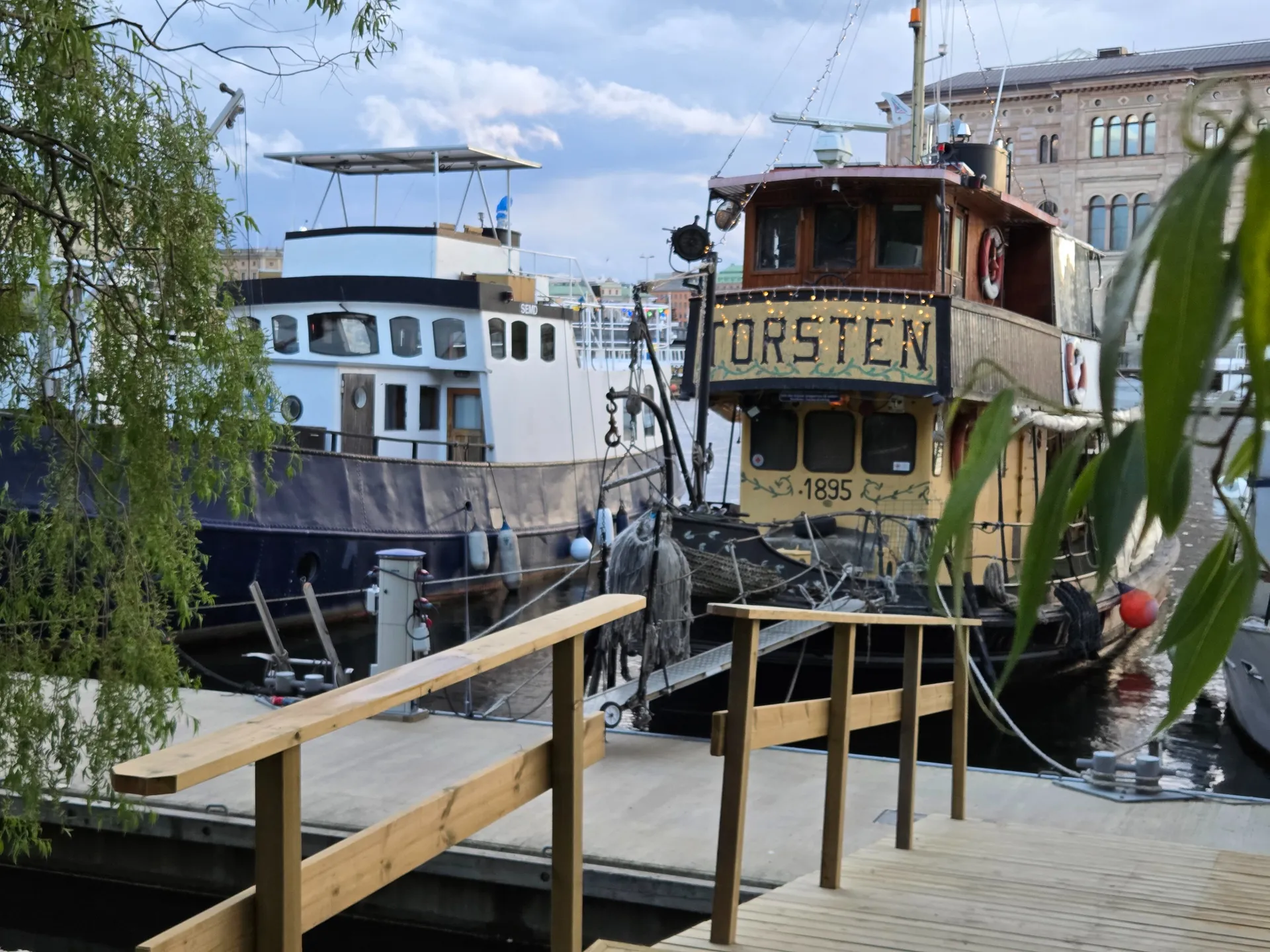 Två båtar vid en träbrygga med räcke. Den ena båten har namnet TORSTEN och årtalet 1895. Gröna löv inramar bilden