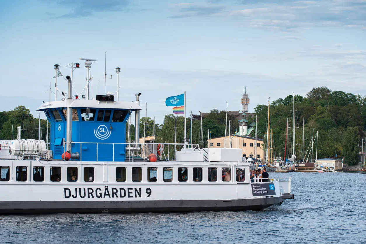 Färjan Djurgården 9 med SL-logga och regnbågsflagga på vattnet. Bakgrund med träd, hus och segelbåtar