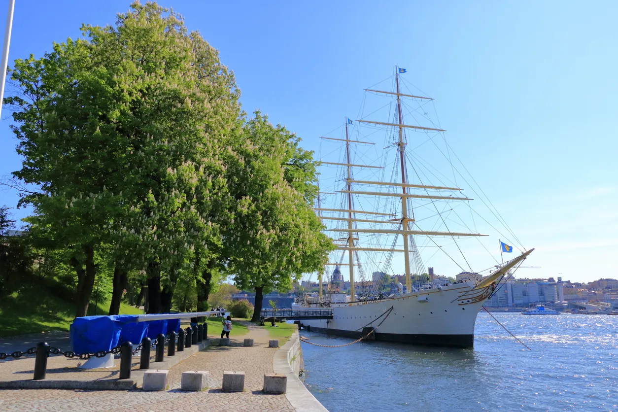 Vitt segelfartyg vid kaj med gröna träd och en stadssilhuett i bakgrunden under klarblå himmel